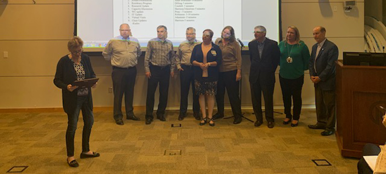 Unmasked team members crowd around a presentation screen and receive their PFAC Award of Excellence before the coronavirus pandemic.