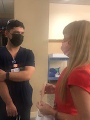 RJ Suarez wears a mask and scrubs while standing next to Zoe Marcus who is wearing a red dress, as she hands him the PFAC Award of Excellence