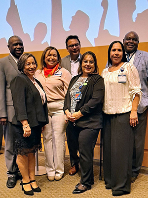Seven people stand together. There are three men in the background and four women in the foreground. They are smiling, professionally dressed and wearing badges.
