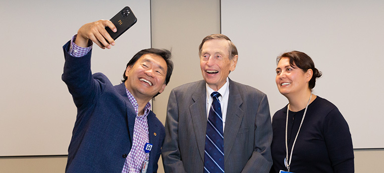 Three people stand together to take a selfie. The man on the left holds his phone out to take the photo while the man in center and the woman on the right smile towards the camera.