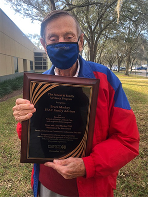 An elderly man in a white and blue windbreaker jacket holds an award plaque and wears a mask while standing outside on a sunny day.. Mackey was the honorary first recipient of the Bruce and Loyce Mackey Patient and Family Advisory Program Volunteer of the Year Award.