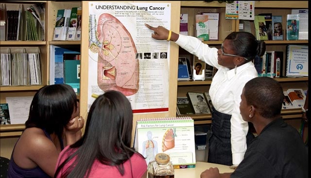 A woman points to a poster about lung cancer while students look on