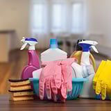 The image shows a collection of cleaning supplies neatly arranged on a wooden surface in a bright and modern home setting. The supplies include various bottles of cleaning sprays, sponges, and a pair of pink rubber gloves draped over a blue plastic basket. 
