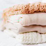 The image displays a neat stack of folded, cozy sweaters in soft, neutral tones, including shades of cream, beige, and light peach.
