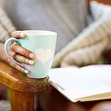 The image shows a close-up of a person holding a light blue mug with a white heart design. The person is seated comfortably in a chair, wrapped in a cozy blanket, and is reading an open book resting on their lap. The setting suggests a warm, relaxed, and peaceful moment.