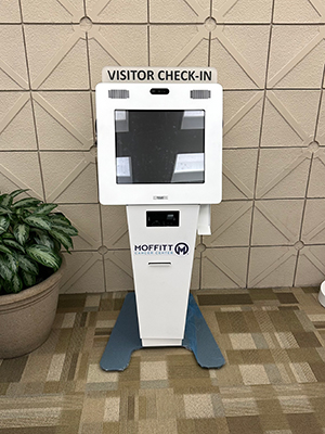 A white electronic kiosk is positioned against a wall. It holds a computer screen and has a sign reading VISITOR CHECK IN across the top of the kiosk. The kiosk stand also has Moffitt Cancer Center's logo on it.