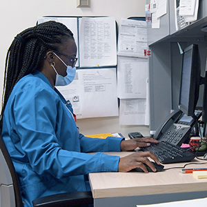 Themika Lewis, a registered nurse, wears blue scrubs and a surgical mask while sitting at her desk and prepares to call a patient. Lewis has black braided hair and wears glasses.