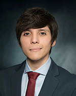 A man wearing a black coat, light blue shirt and dark red tie smiles gently while posing in front of a dark blue photo backdrop. He has lighter skin tone, dark black hair and dark brown eyes. Dr. Luis E. Aguirre, Malignant Hematology Program