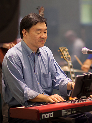 Dr. Patrick Hwu looks down while playing a keyboard and wearing a light blue shirt with rolled up sleeves.