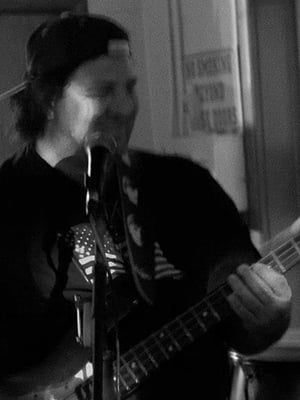 A black and white image shows Jeff Leighton wearing a backwards baseball cap while playing bass.