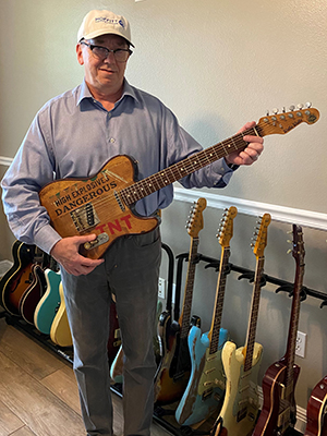 Dr. James Mule wears a gray shirt while holding a guitar that has TNT High Explosives written across it. He stands in front of his collection of guitars.