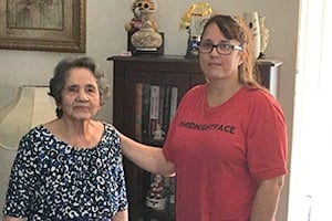 Edna, wearing glasses and a red shirt, is standing beside her mother who is wearing a floral pattern shirt. Edna has her hand on her mother's shoulder. Both are smiling.
