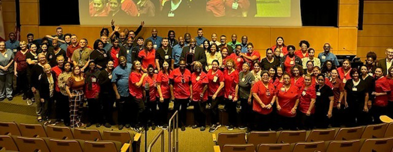 More than a hundred people gather in Moffitt's auditorium for a photo. They're standing on the stage in front of a presentation screen while wearing their blue and red uniforms.