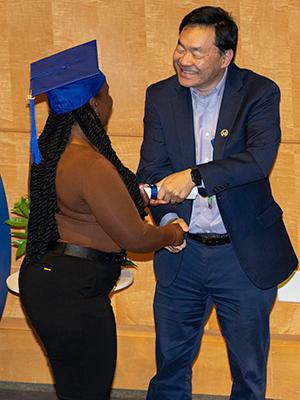 A black woman with long braids wears a blue graduation cap while shaking a man's hand. The man is handing her a diploma and smiling. The face of the Black woman can't be seen in the photo.